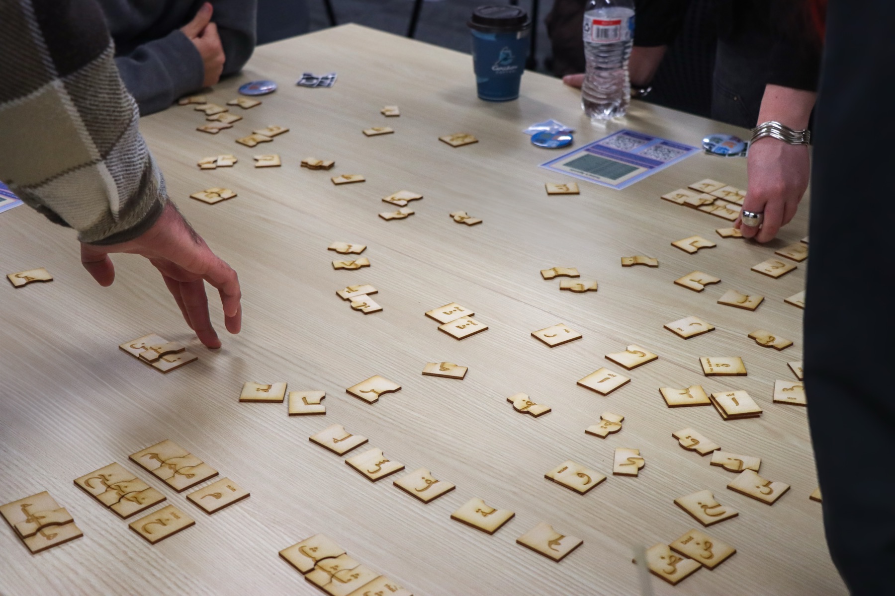 Hands reaching across a table covered in scattered Huruf La'b wooden letter tiles during a game.