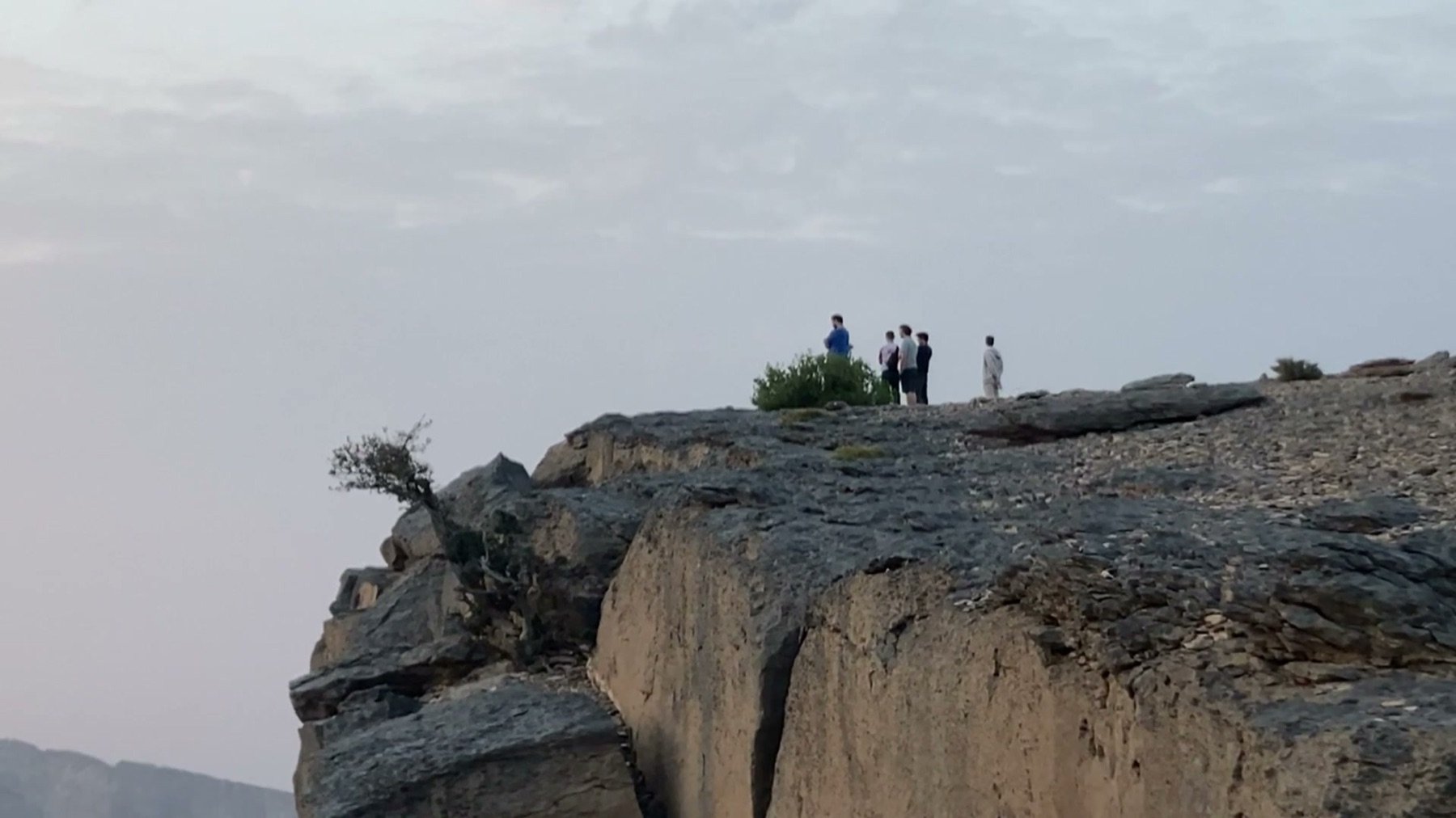A small group stands on the overhanging rim of Jebel Shams at dusk — the highest peak in Oman — above the haze of the Wadi Ghul canyon.
